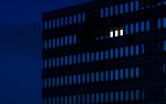 A dark building at night, the black building structure contrasting with a deep blue night sky, with many vertical rectangular windows, three windows are brightly lit.