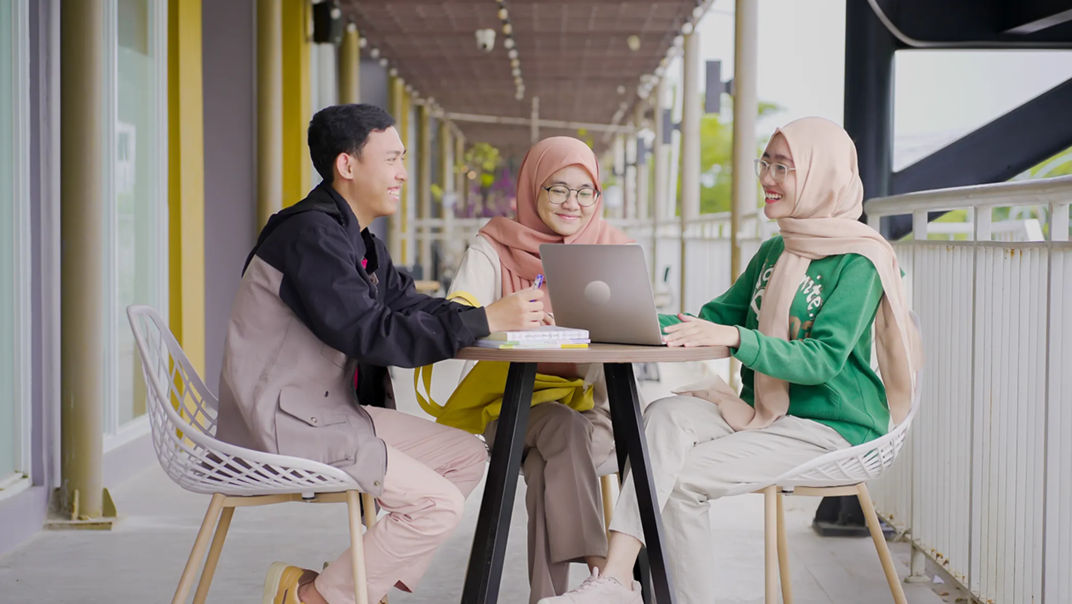 A man wearing a jacket and two women wearing pink headscarves sit at a table outside, smiling. A laptop and writing materials are on the table.